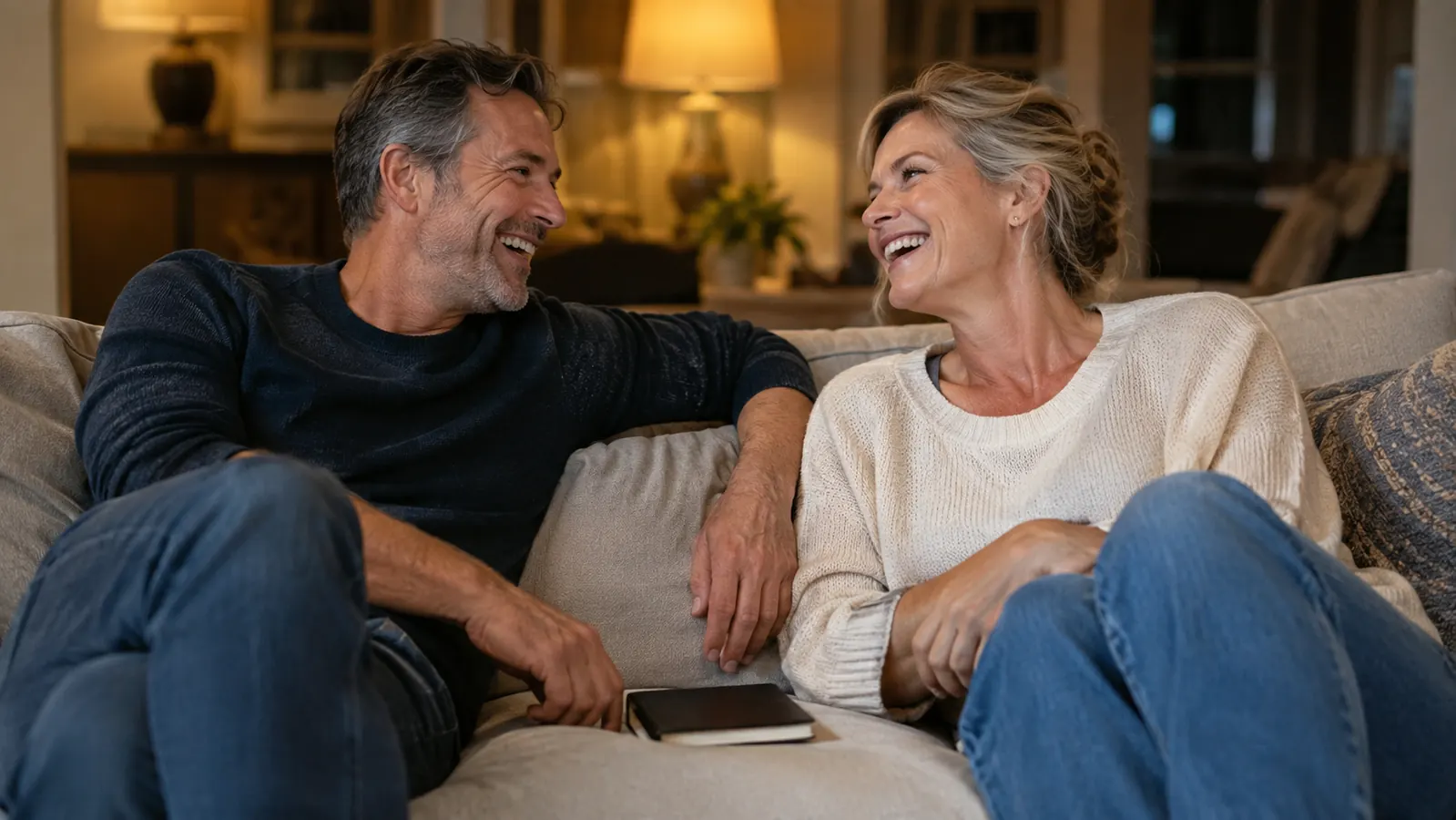 A couple laughing together on a couch in the evening, a journal resting between them